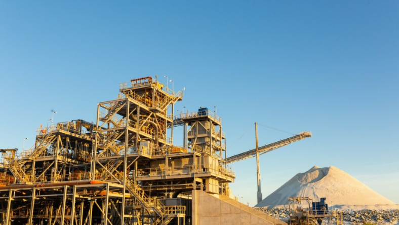 Industrial processing facility with conveyors and metal structures beside a large mineral stockpile